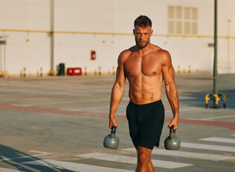 Muscular,Athlete,Walking,And,Carrying,Two,Kettlebells,In,A,Parking